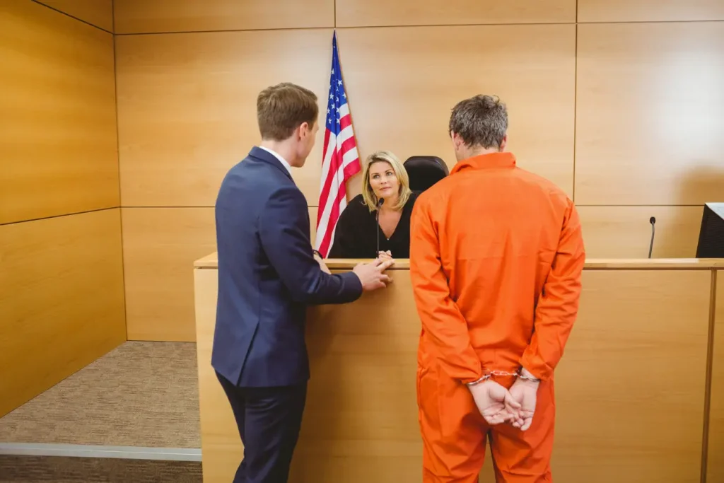 Criminal defense lawyer and handcuffed inmate in an orange jumpsuit standing in front of a female judge in court.
