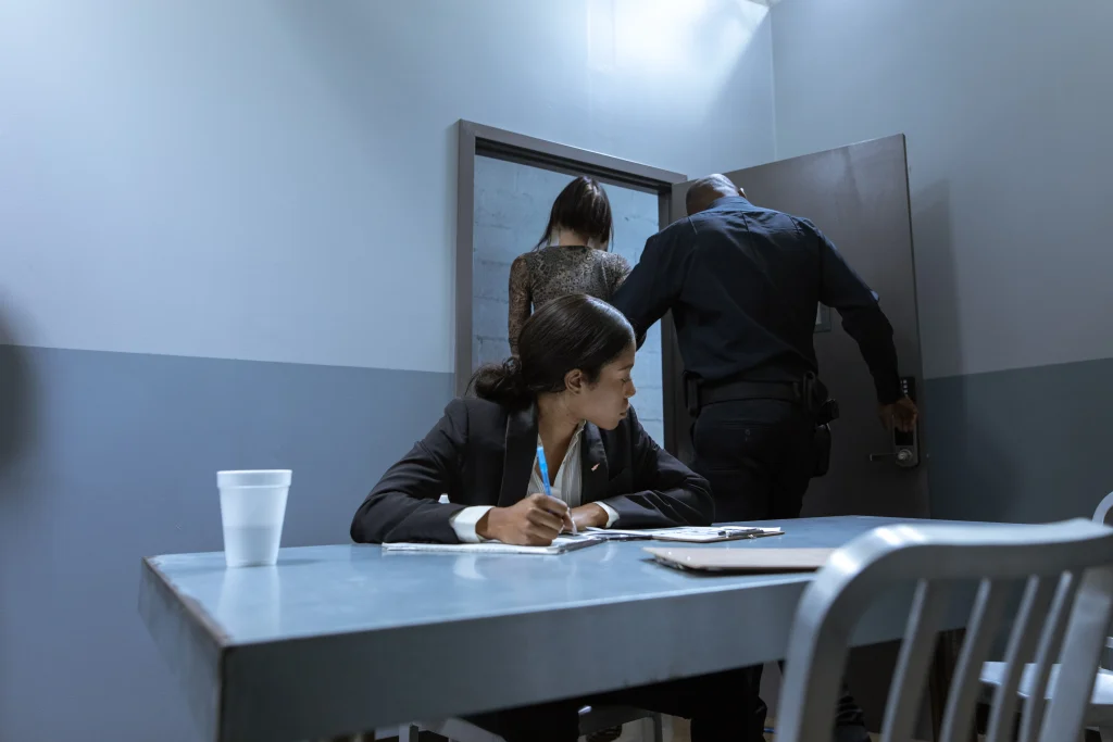A woman being escorted out of an interrogation room by a cop with a lawyer sitting at the table